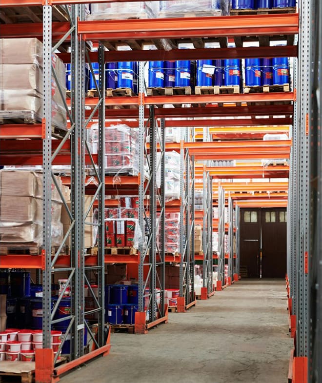 Chemical storage facility with tall steel shelving racks holding pallets of boxes, containers, and barrels, organized along wide concrete aisles
